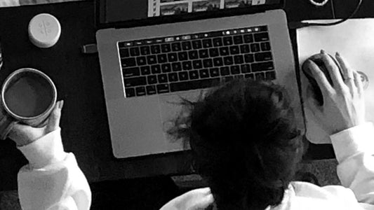 This black and white overhead view of a girl, hair up in a bun, wearing a white hooded sweatshirt, sitting at her silver computer, hand on the mouse, other hand holding her coffee cup.
