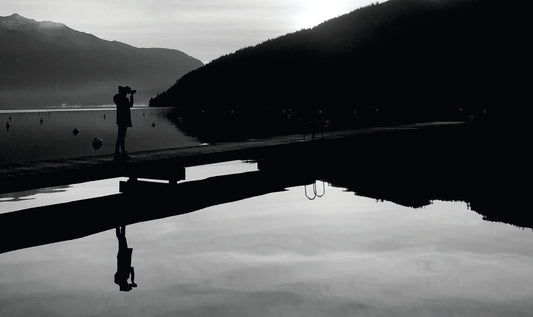 The black and white image depicts a black silhouette of a photographer standing on a dock and aiming her camera at a mountain. The silhouette scene and lighter cloudy sky is reflected in the silver lake.