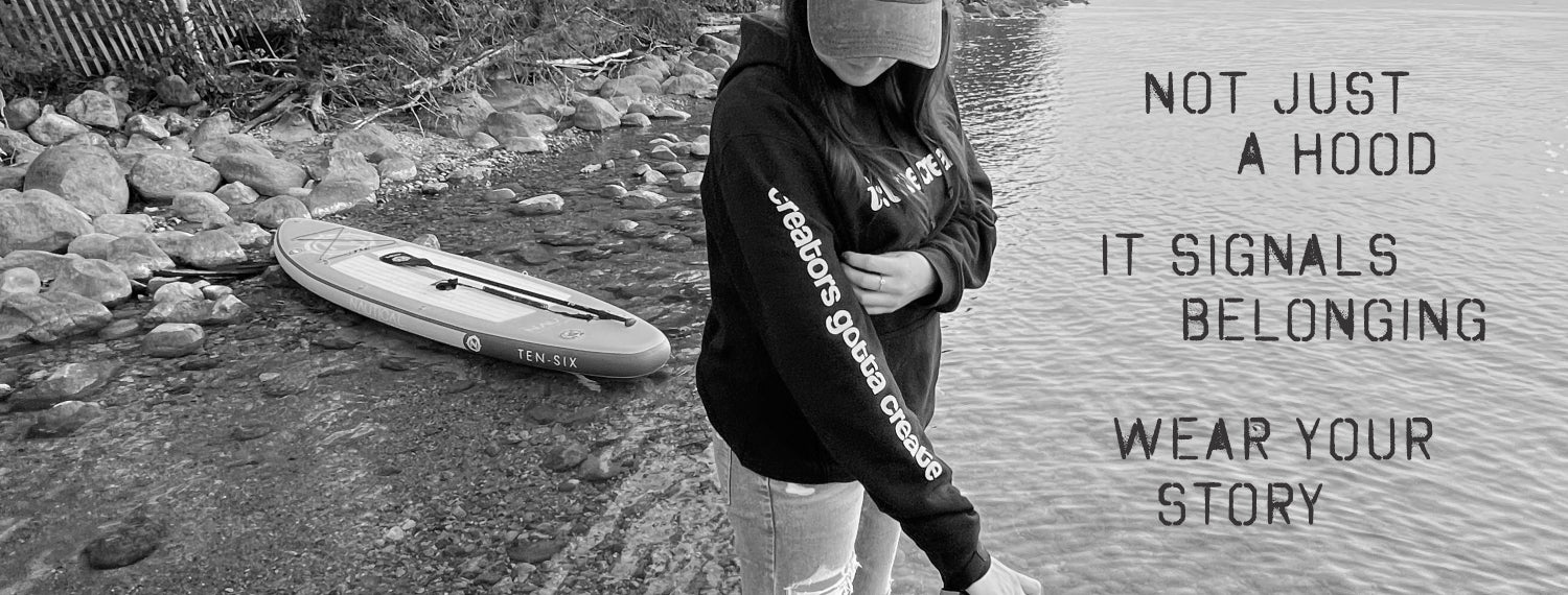 A black and white lifestyle photograph of a person wearing a black hoodie with 'urban outdoor gear' printed on the sleeve, standing next to a paddleboard on a rocky shoreline with water on the right. Text on the right reads 'NOT JUST A HOOD IT SIGNALS BELONGING WEAR YOUR STORY'.